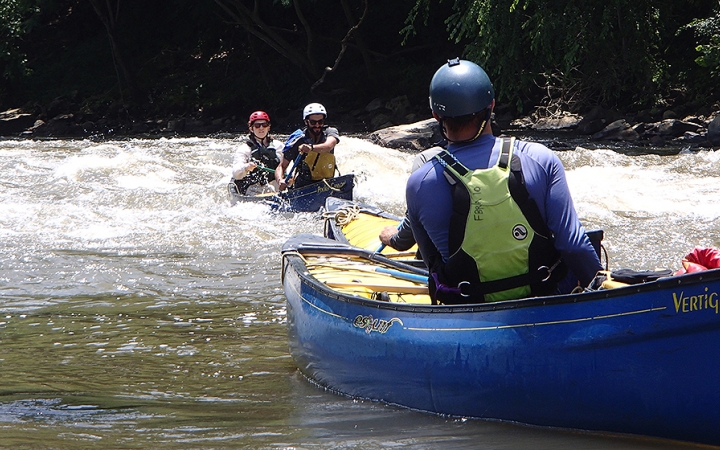 Two students paddle a canoe through whitewater, while another person who is likely an instructor watches them from another canoe.
